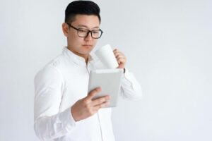 안경쓴 동양인 남성이 차를 마시며 타블렛을 보고 있다.